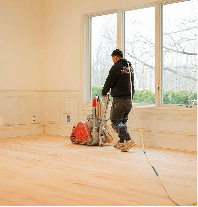Floor refinisher operating a drum sander on hardwood floors in a residential room